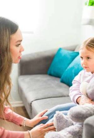 Caucasian female psychologist talking and comforting a sad little girl during a child therapy session. Elementary girl hugging a teddy bear and listening to her therapist