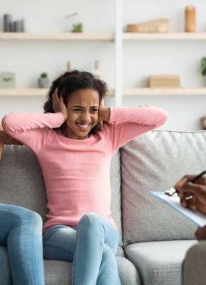 Black teen girl with behavioral disorder having therapy session with child psychologist, sitting on couch next to mother, covering ears, feeling annoyed while having conversation with psychotherapist